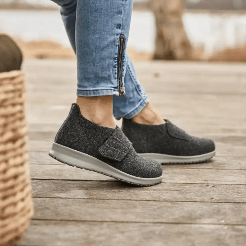 Vrouw draagt antracietgrijze New Feet pantoffels op een houten vloer, zijaanzicht van de voeten
