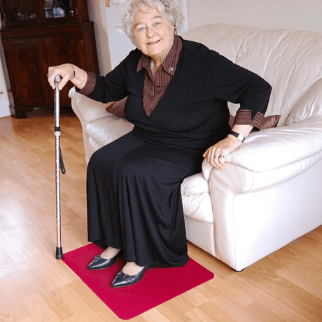 Femme âgée assise sur un canapé avec une canne et les pieds sur un tapis antidérapant rouge