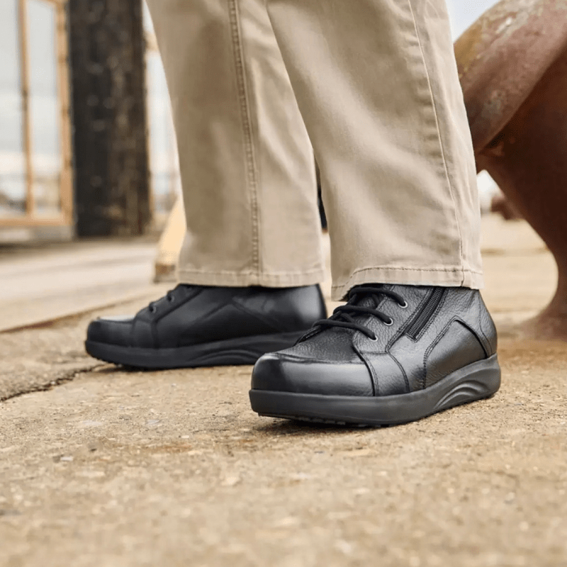 Een man in beige broek en zwarte New Feet herenboots staat op een grijze ondergrond