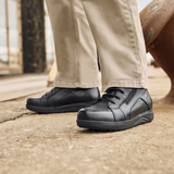 Een man in beige broek en zwarte New Feet herenboots staat op een grijze ondergrond